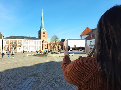 Bild vergrößern: Frau mit Tablet sieht den heutigen Markplatz; Auf dem Tablet ist der historischer Marktplatz zu sehen.